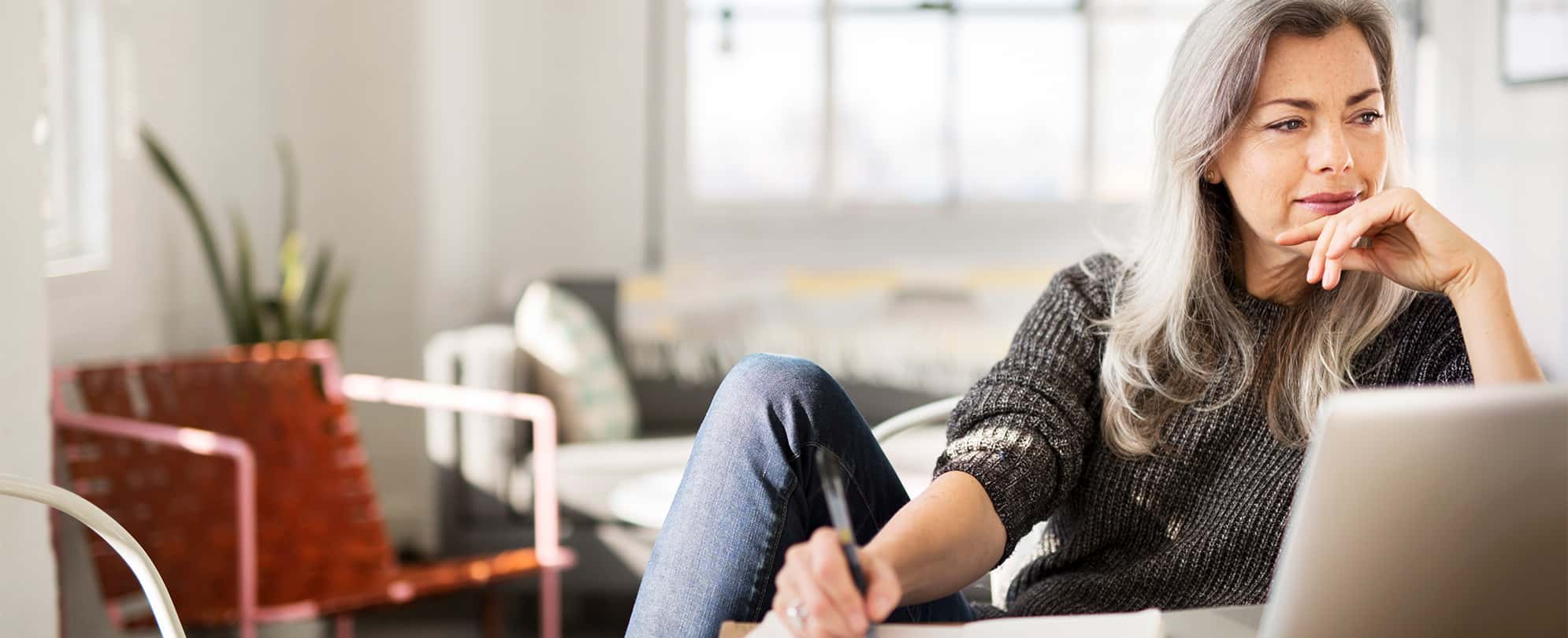 A woman sitting in front of a laptop while writing on a piece of paper, stares off into the distance thoughtfully.