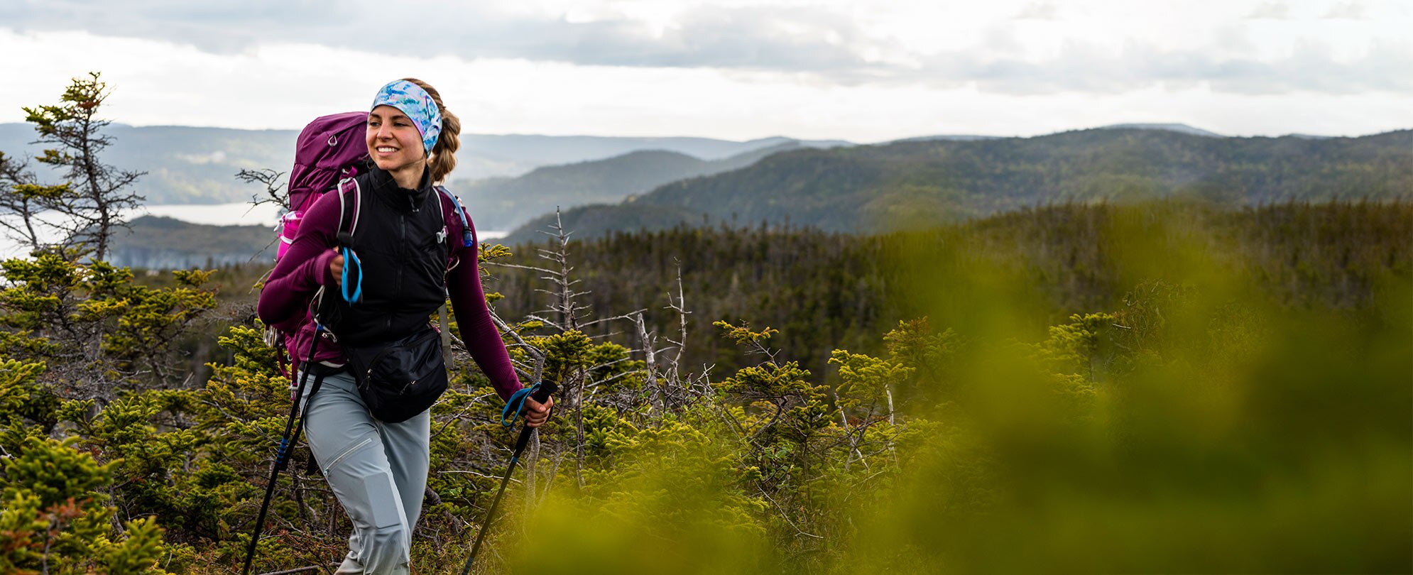 A woman wearing a headband and backpack uses trekking poles to hike in the mountains of a National Park.