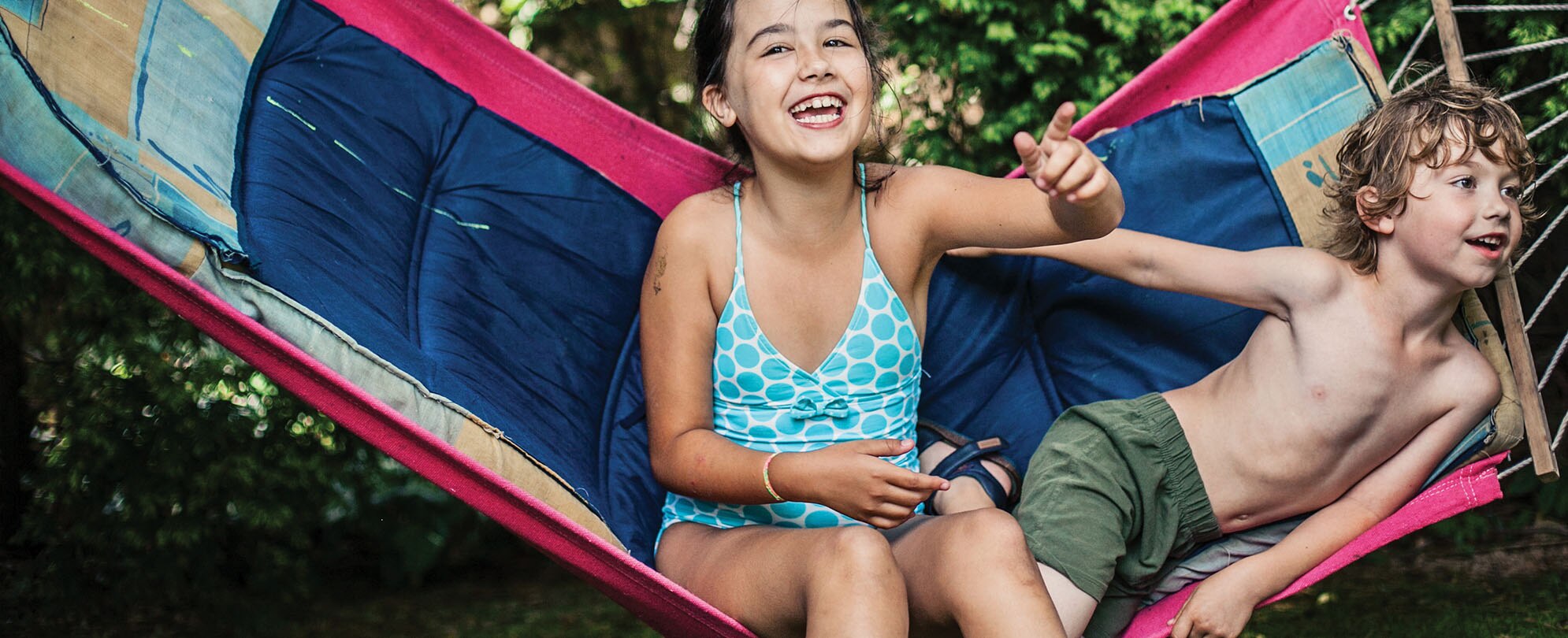 A young girl and boy wearing swimsuits sit in a hammock.