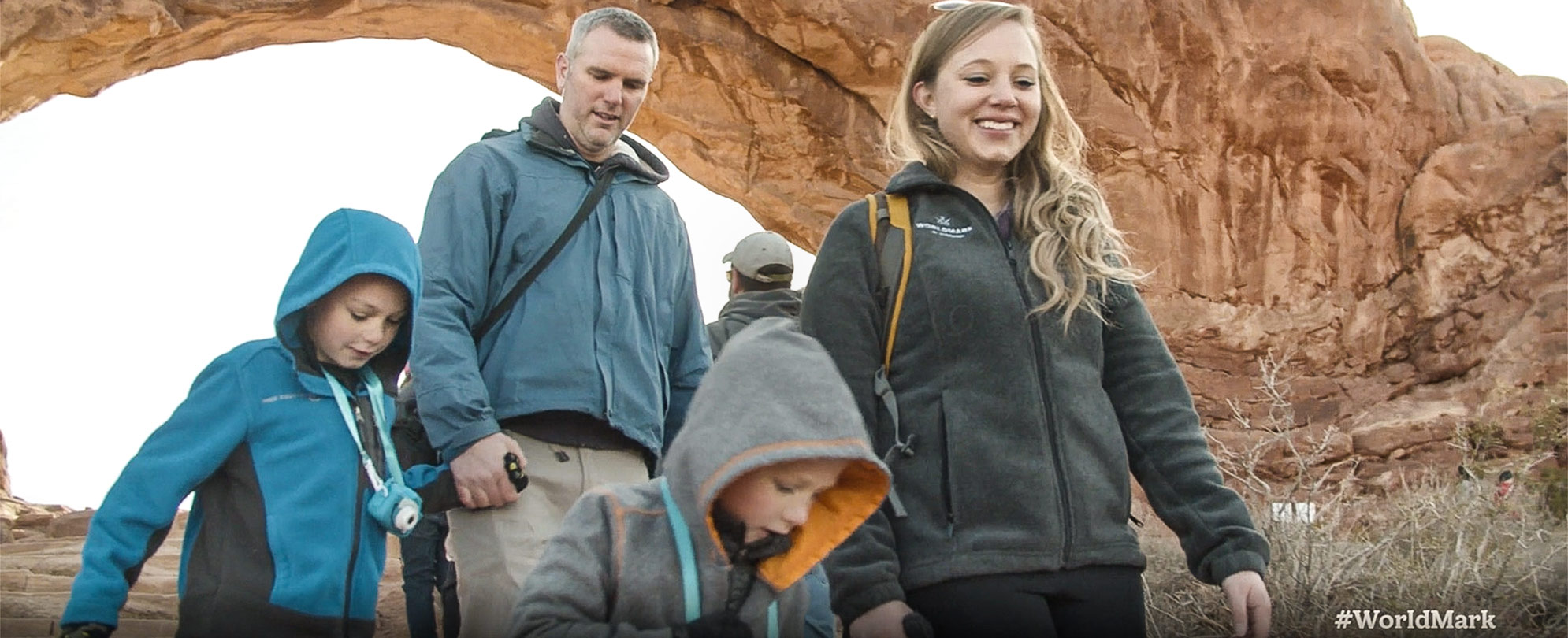 Woman smiles while hiking down a hill with a man walking behind her carrying a young girl on his shoulders and pointing to something out in the distance.