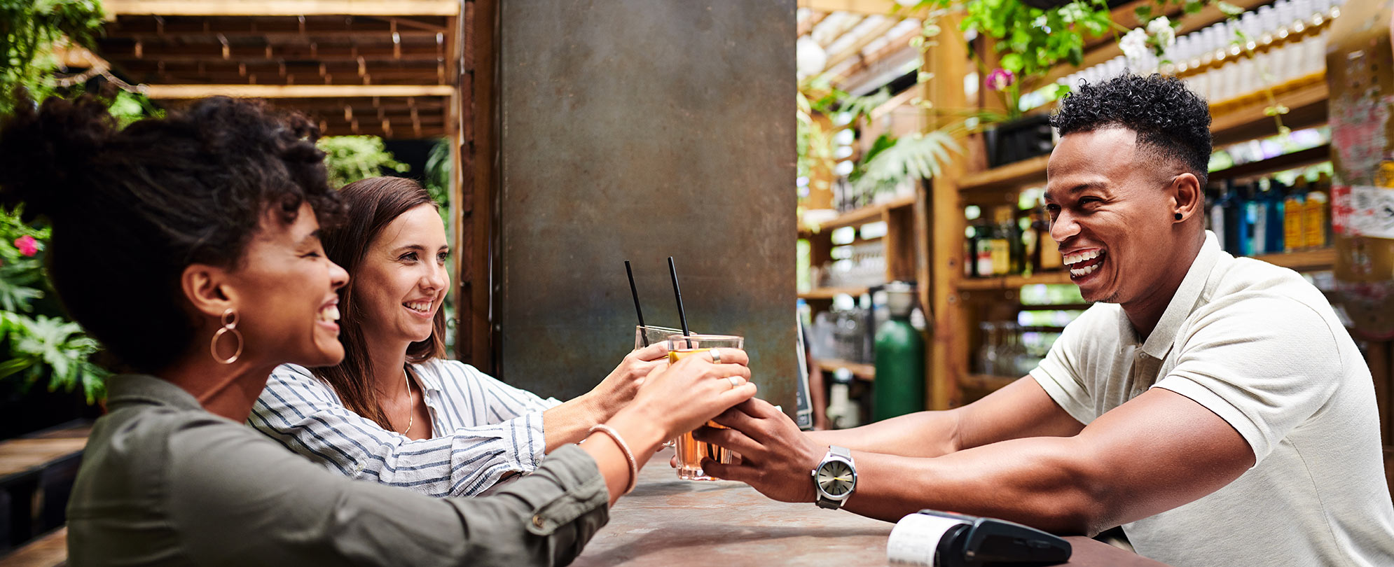 Smiling man serving two smiling women drinks over a bar.