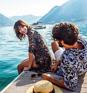 A man and woman sitting on a dock by the lake with mountains in the distance. 